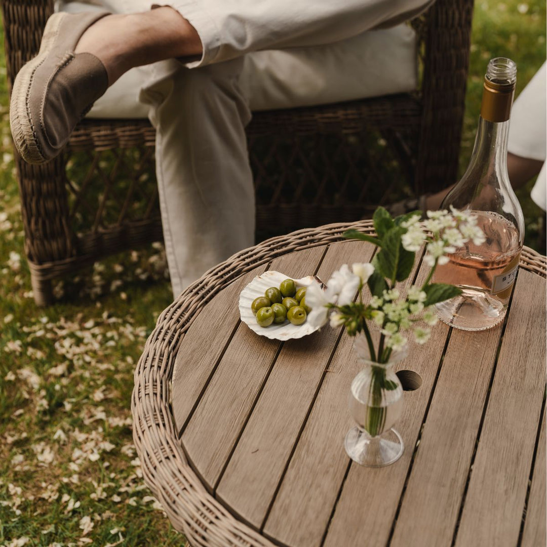 Person sitting outdoors with a wooden table featuring a bottle, flowers, and fruit.