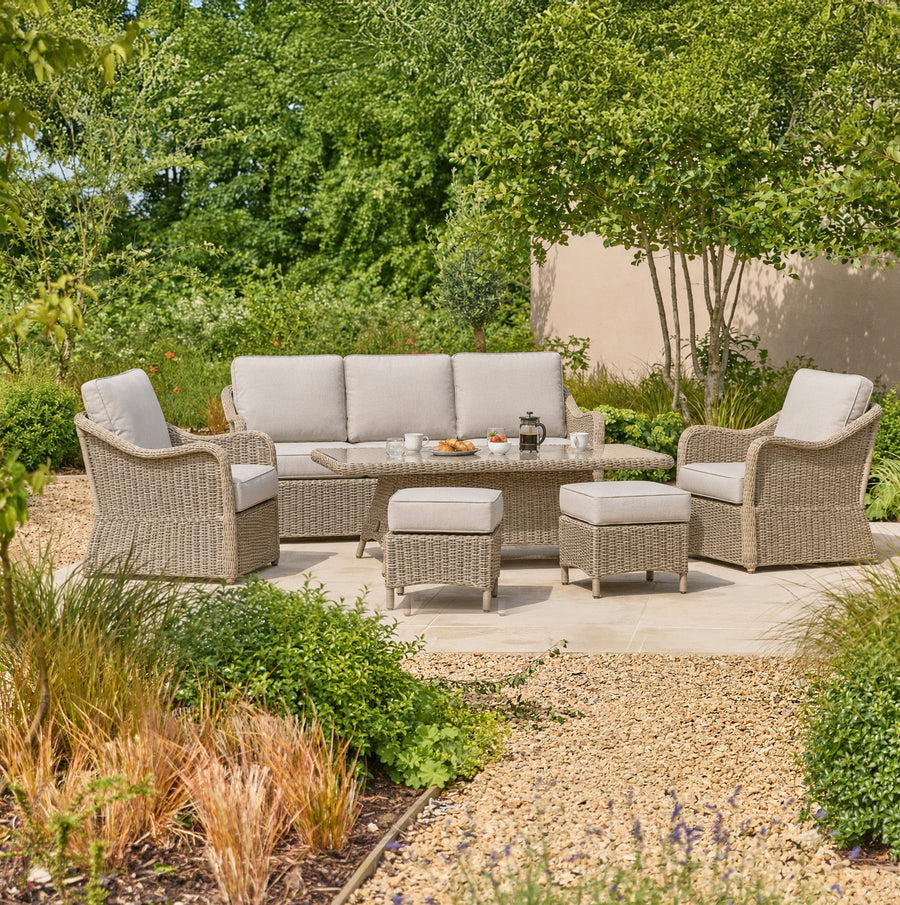 A 7-seat outdoor patio furniture set made of warm grey woven wicker, featuring a 3-seater sofa, 2 armchairs, a height adjustable table, and two ottomans with light gray cushions, arranged on a stone patio in a landscaped garden.