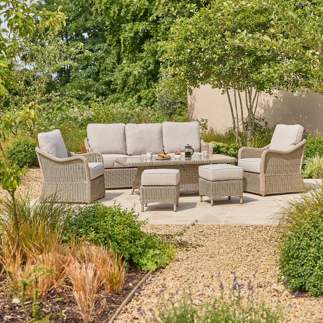 A 7-piece outdoor patio furniture set made of warm grey woven wicker, featuring a 3-seater sofa, 2 armchairs, a height adjustable table, and two ottomans with light gray cushions, arranged on a stone patio in a landscaped garden.