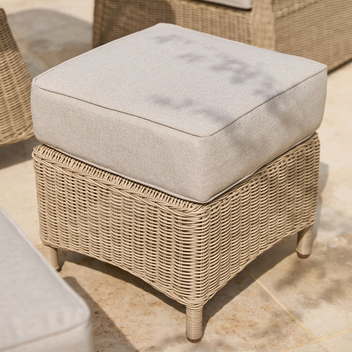 A square outdoor wicker ottoman with a thick, light beige textured cushion on a tiled patio.