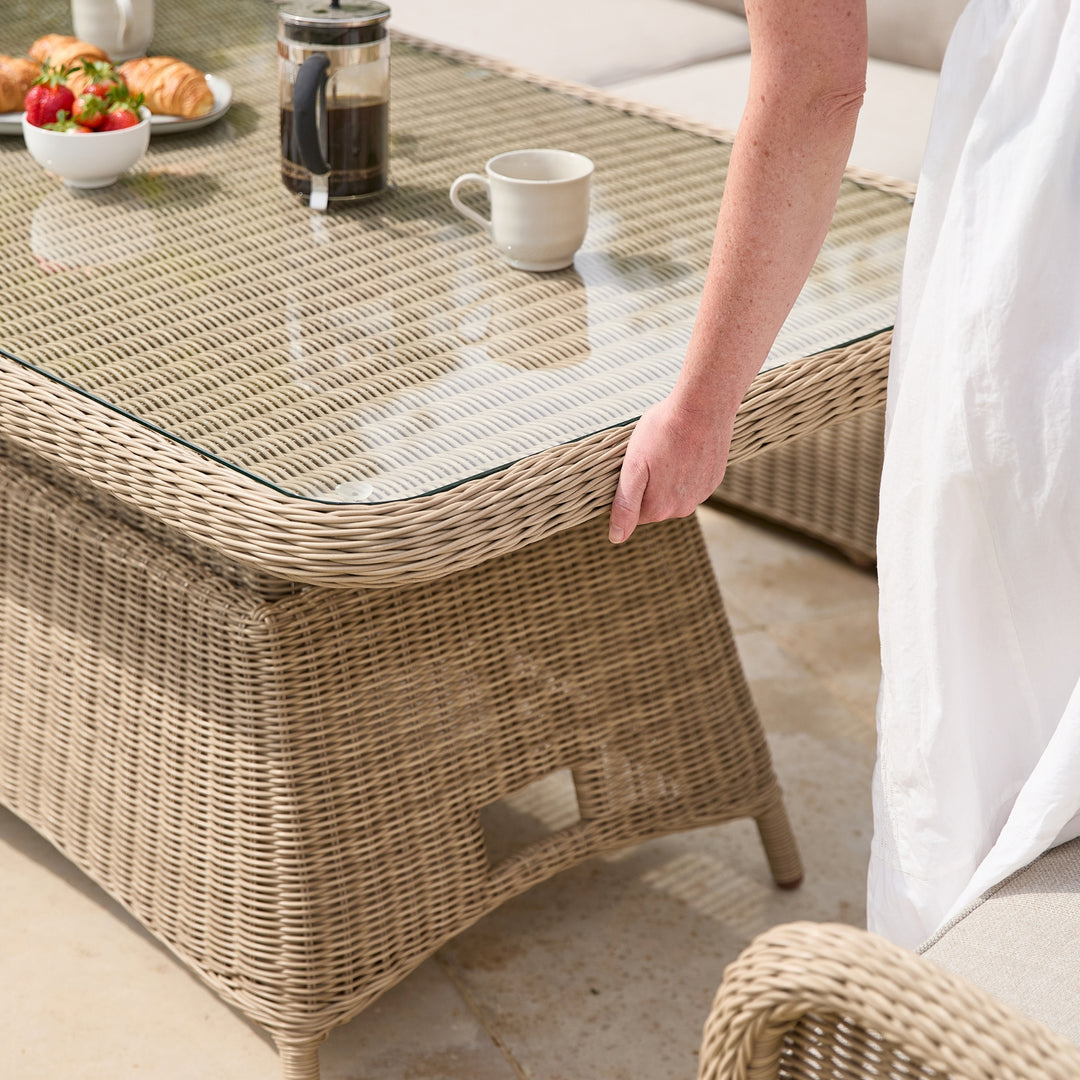 A woven rattan outdoor coffee table with a clear glass top, shown in a patio setting with a person demonstrating the height adjustable table