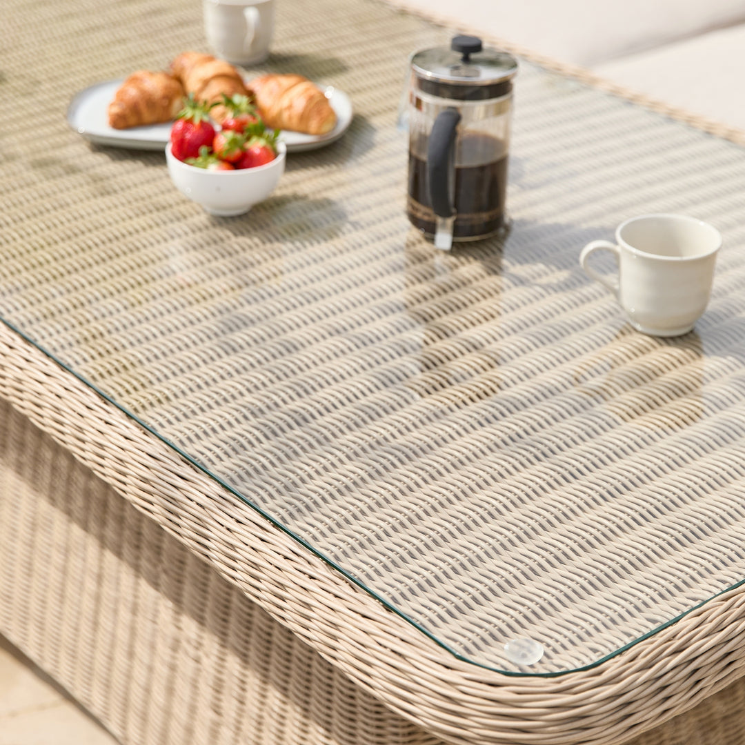 A woven outdoor dining table with a clear glass top, styled with a plate of croissants, a bowl of strawberries, a coffee press, and a white mug in a bright patio setting.