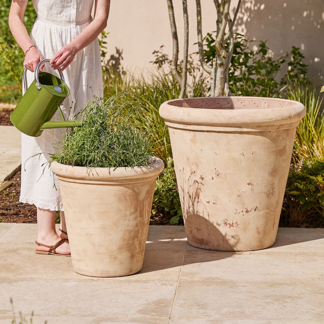 Person watering a plant in a large pot with another pot nearby on a patio.