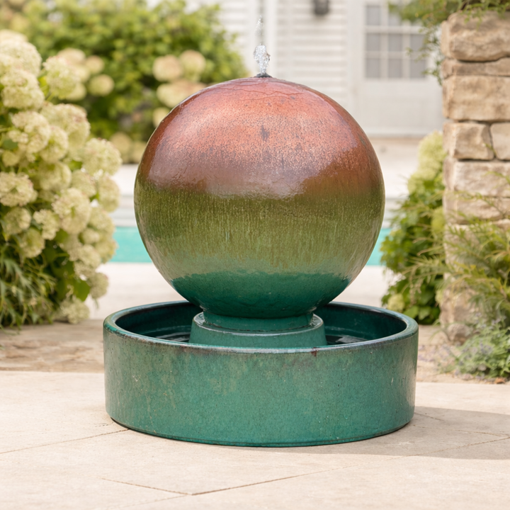 Extra-large globe-shaped ceramic water feature with a aqua bronze verdigris glaze and integrated basin, styled on a garden patio.