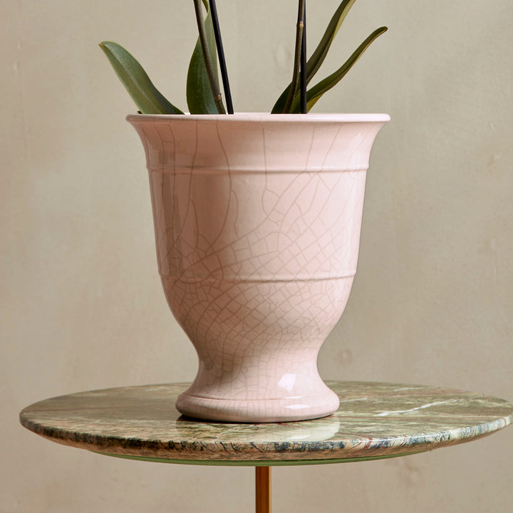 Pink ceramic urn plant pot with a cracked texture on a round marble table against a beige wall.