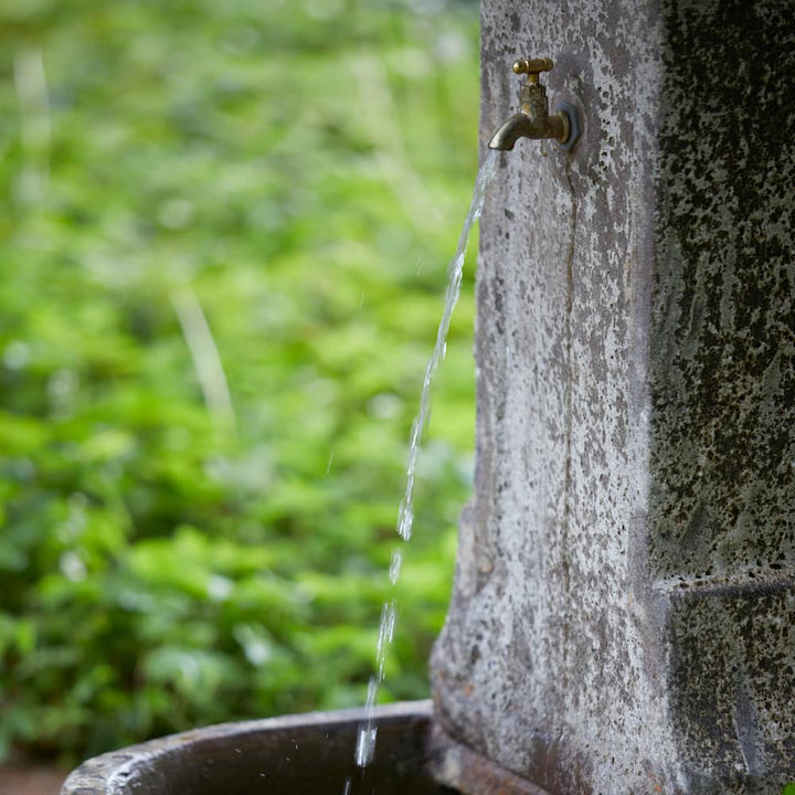 Ancient stone garden fountain in action, with water gently flowing from the brass tap into the weathered stone basin.