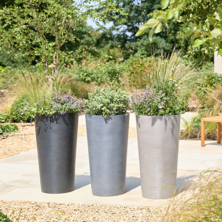 Three tall cylindrical outdoor planters in dark charcoal, medium gray, and light gray, filled with ornamental grass and small purple flowers, arranged on a concrete patio in a landscaped garden.