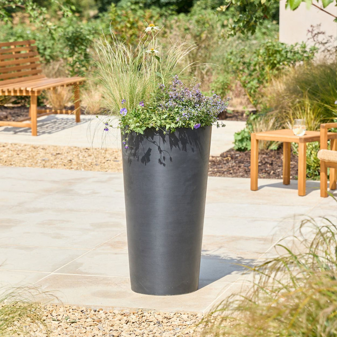 A tall, dark charcoal gray cylindrical planter filled with ornamental grass and small purple flowers, set on a light stone patio in a landscaped garden with wooden benches and tables.