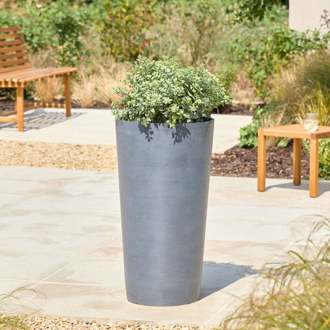 A tall, cylindrical dark gray planter filled with green foliage, set on a light stone patio in an outdoor garden setting with wooden furniture in the background.