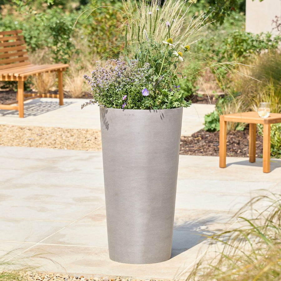 A tall, cylindrical concrete planter in a light gray finish, filled with ornamental grasses and small purple flowers, set on a stone patio in a landscaped garden with wooden benches and a side table.