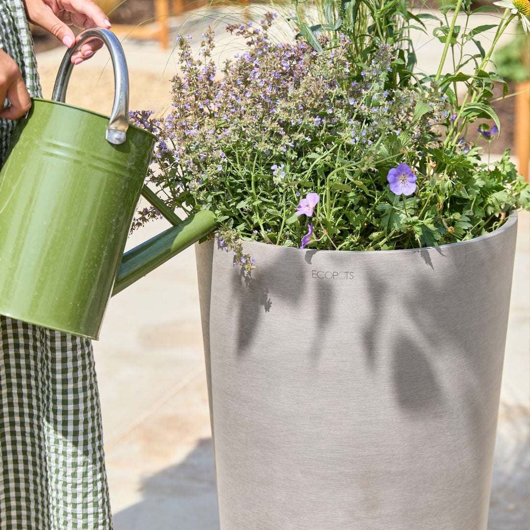A person in a green-and-white checkered dress holds a green metal watering can beside a large light gray cylindrical planter filled with green plants and small purple flowers in an outdoor setting.