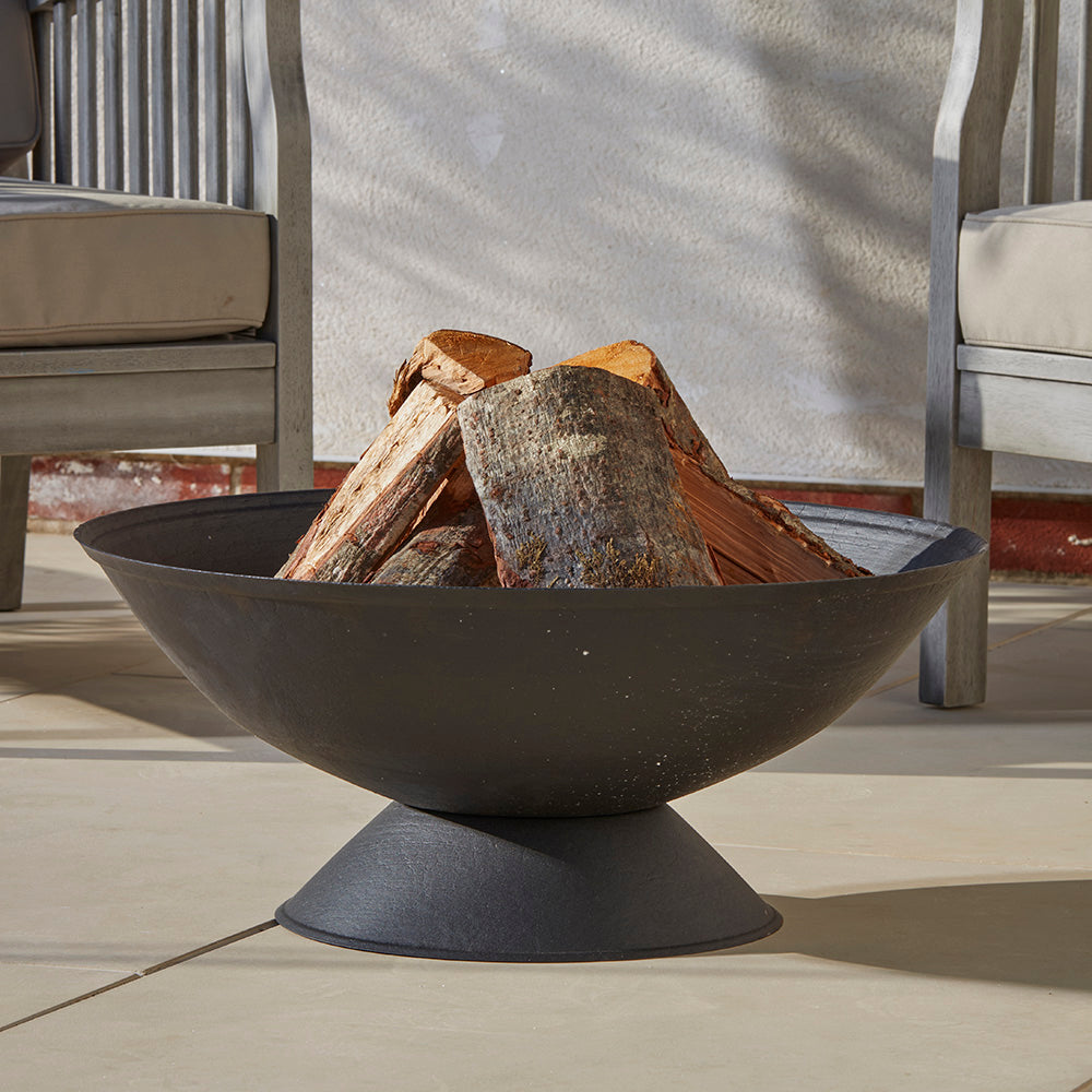 A black cast iron fire pit with logs stacked, ready to light when the sun goes down, placed on a buff coloured patio, creating a cozy outdoor atmosphere.