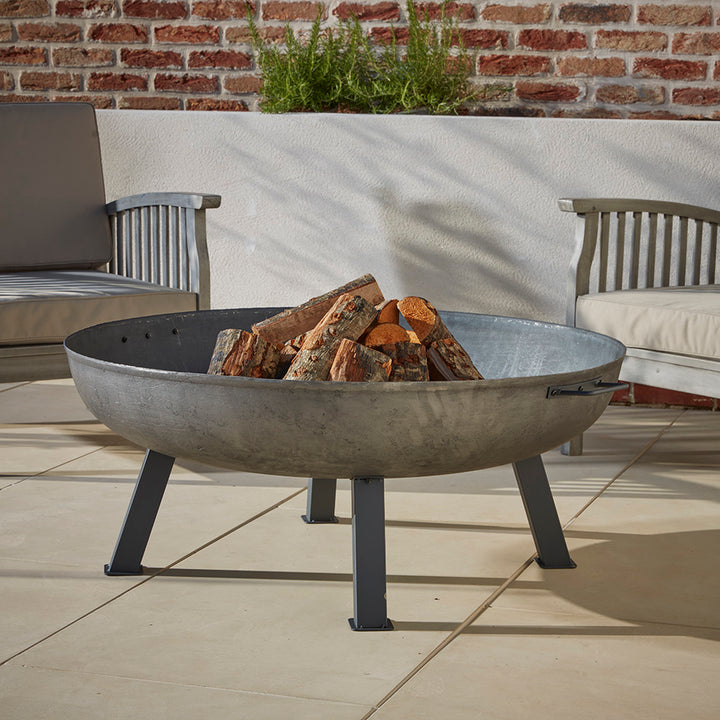 A cast iron Fire Pit Bowl stacked with logs ready to be lit on a cool evening in the garden. Nestled in-between two wooden garden chairs