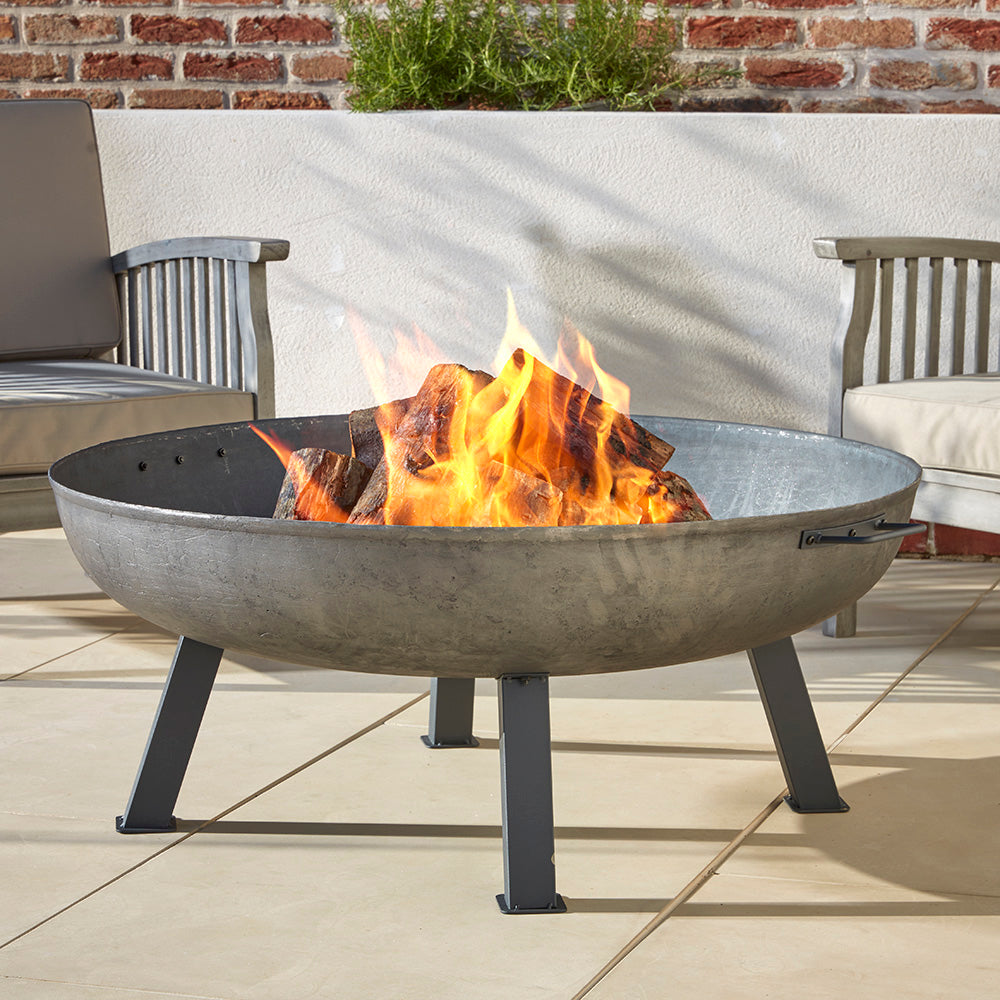 Hoole Cast Iron Fire Pit Bowl with a roaring fire, placed on a stone patio. A durable, traditional fire pit with sturdy steel legs, perfect for garden gatherings.