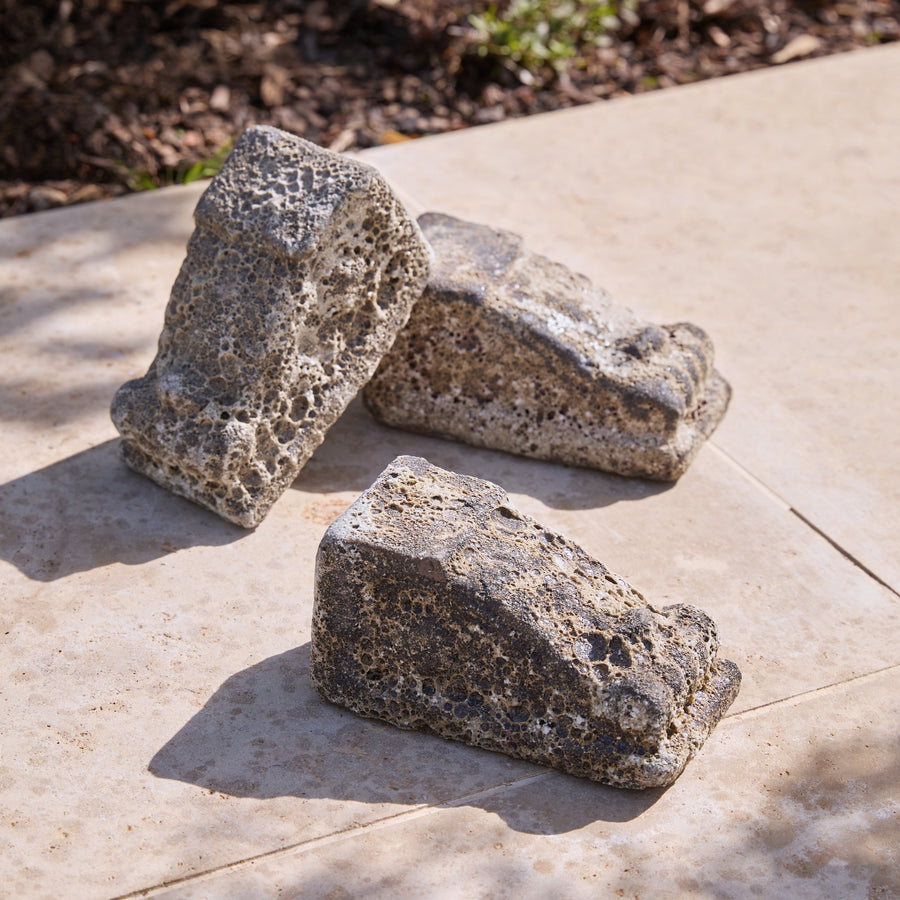 Three rustic porous stone garden pot feet in an ancient salt glaze finish, arranged on a light beige stone patio with outdoor mulch visible in the background