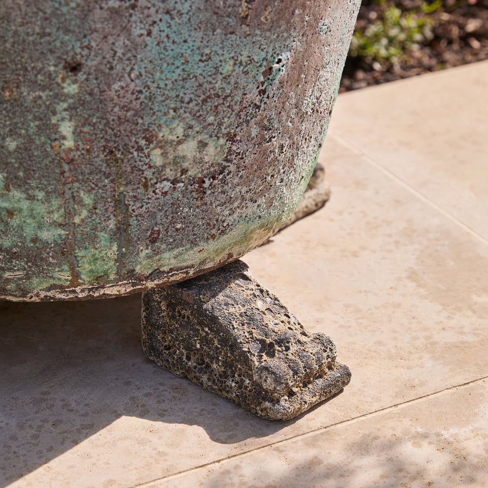 A close-up outdoor shot of a large weathered stone or metal planter pot resting on a decorative stone base, set on a light beige tiled patio.