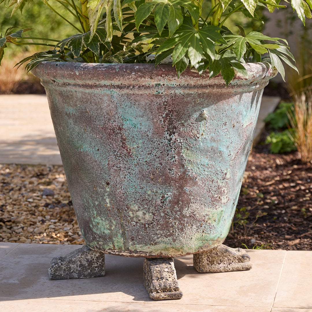 A large decorative outdoor planter with a distressed turquoise and brown patina finish, featuring three sculpted feet and filled with green foliage, set on a patio in a garden.