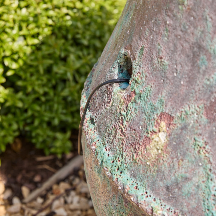 Close-up of a textured stone surface with a wire showing where the water feature cable comes from, against a blurred green bush background.