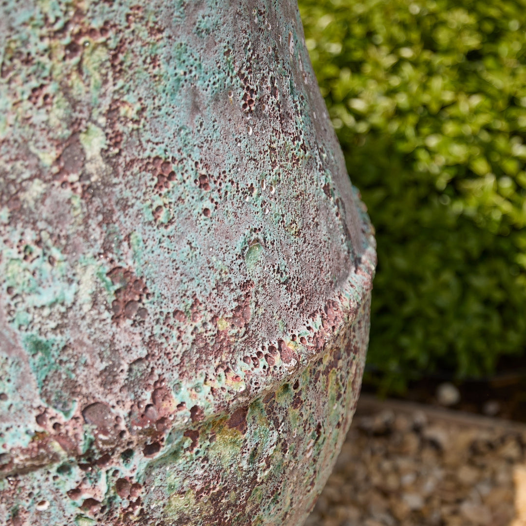 Close-up of a textured verdigris surface with green and brown patina, blurred green foliage in the background.