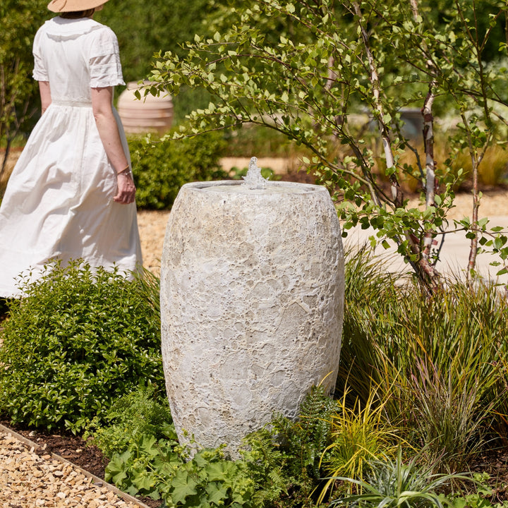 Woman in a white dress standing next to a stone fountain in a garden setting to show how large the water feature is