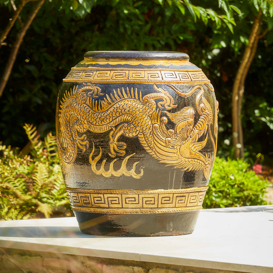Large antique-style Chinese garden pot with detailed dragon motif, placed outdoors on a stone surface surrounded by greenery.