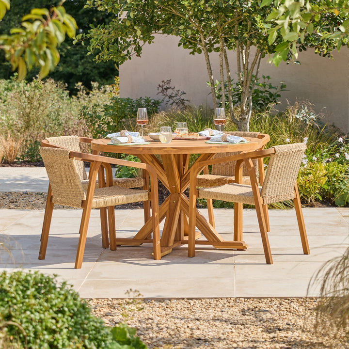 A round wooden dining table with four woven wicker chairs, set on a light stone patio in a landscaped garden with trees and plants.
