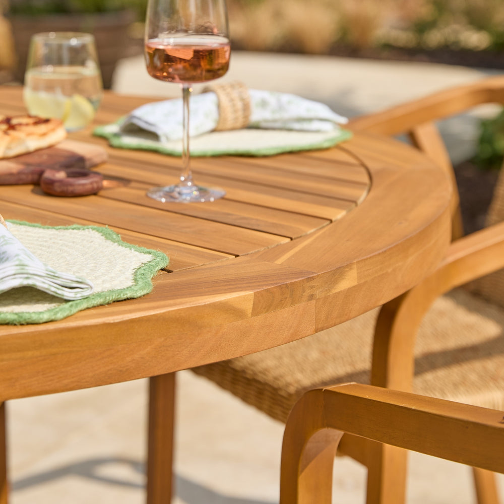 A close-up outdoor shot of a round wooden dining table set with woven chairs, green placemats, folded napkins, and wine glasses on a patio.