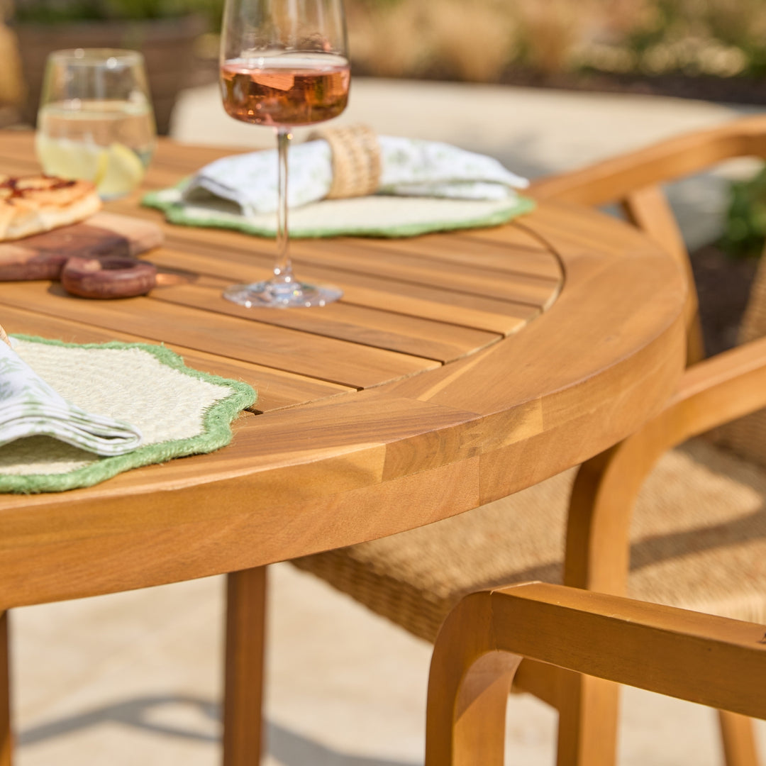 A close-up outdoor shot of a round wooden dining table set with woven chairs, green placemats, folded napkins, and wine glasses on a patio.