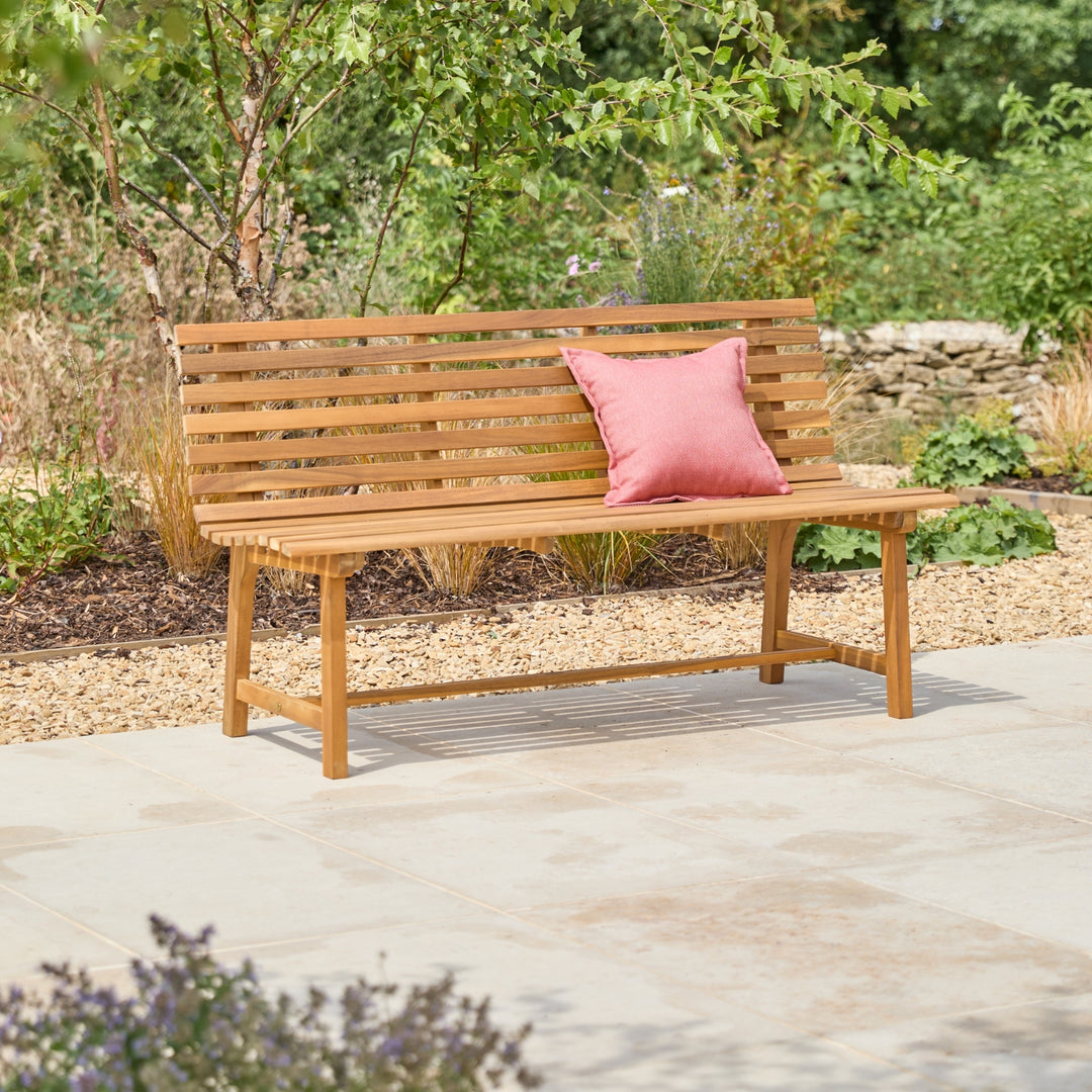 A wooden outdoor bench with a slatted back and seat, placed on a light stone patio with a pink cushion, set against a garden background.
