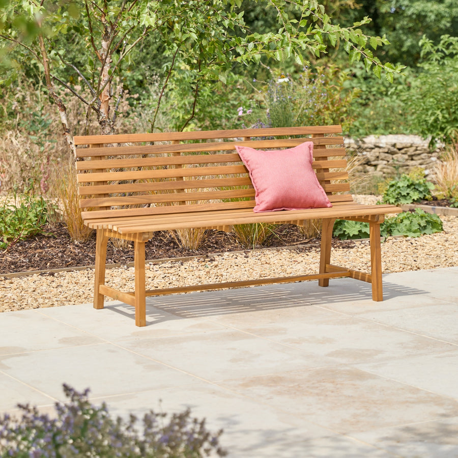 A wooden outdoor bench with a slatted back and seat, placed on a light stone patio with a pink cushion, set against a garden background.