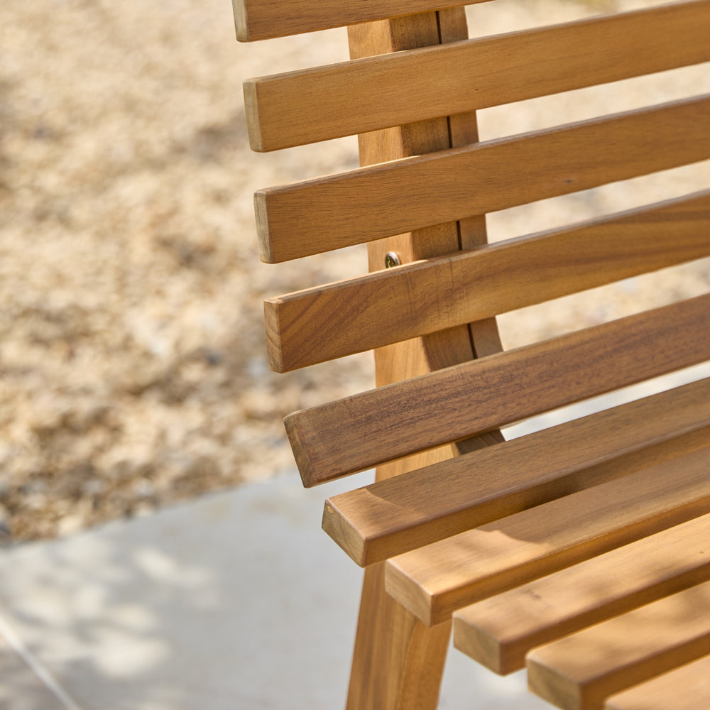 Close-up of a wooden slatted bench or chair outdoors, showing the warm medium-brown wood grain and slatted back and seat, with a blurred gravel and patio background.