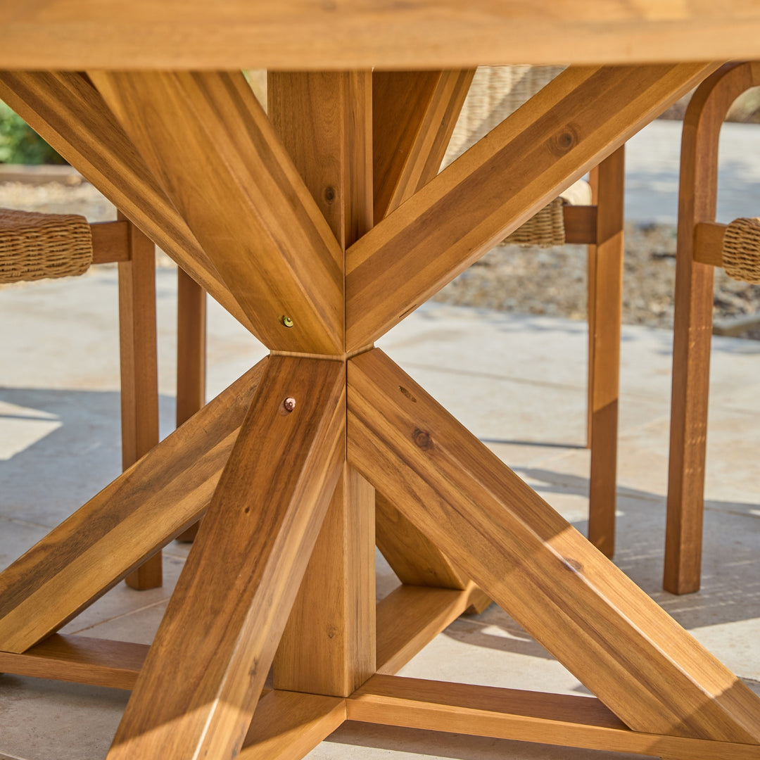 Close-up of a wooden outdoor dining table base with a geometric considered design, showing natural wood grain and metal fasteners, with part of a woven chair visible in the background.