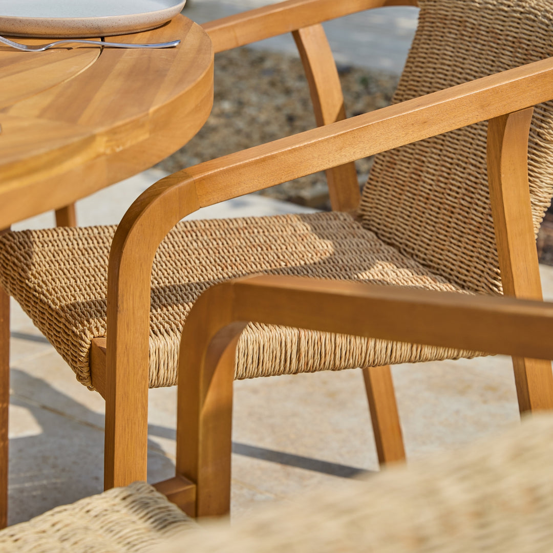 Close-up of an outdoor patio chair with a light brown wooden frame and woven natural fiber seat and back, shown beside a matching round wooden table on a stone patio.
