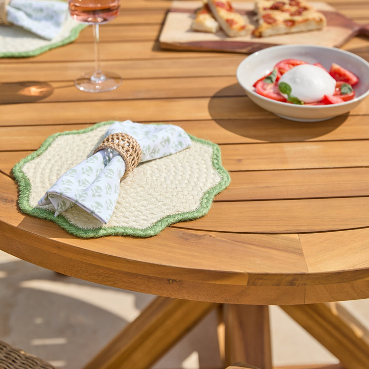 A round wooden outdoor dining table styled with a green and cream scalloped placemat, a patterned napkin in a woven ring, a wine glass, and a bowl of strawberry dessert in the background.