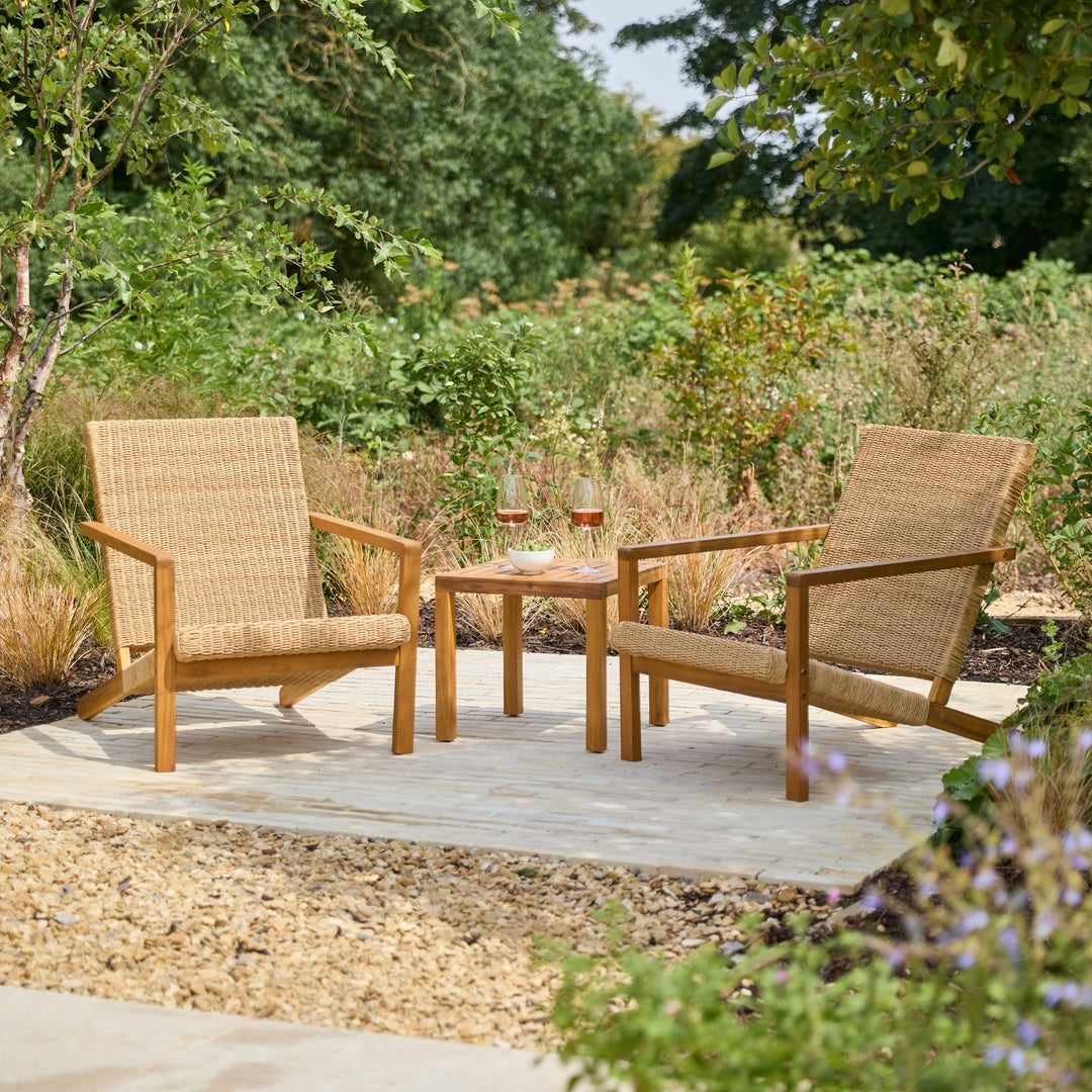 Two woven outdoor armchairs with a small square wooden side table between them, set on a patio in a landscaped garden with greenery and plants in the background.