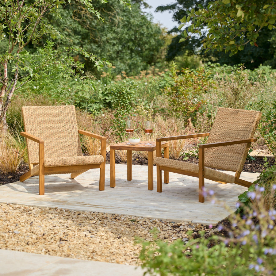 Two woven outdoor armchairs with a small square wooden side table between them, set on a patio in a landscaped garden with greenery and plants in the background.