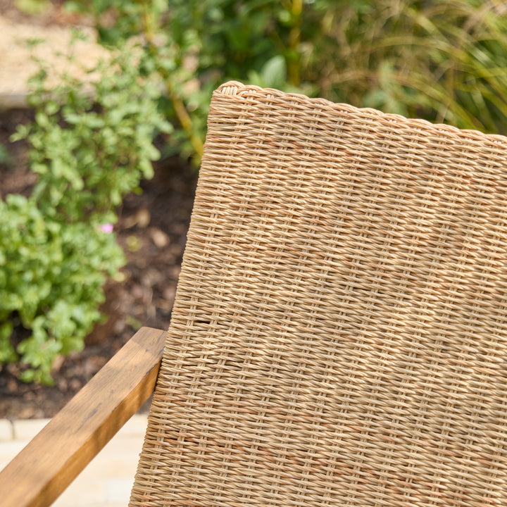 Close-up of a woven wicker or rattan chair back in a natural tan color, with a wooden armrest visible on the left and a blurred garden background.