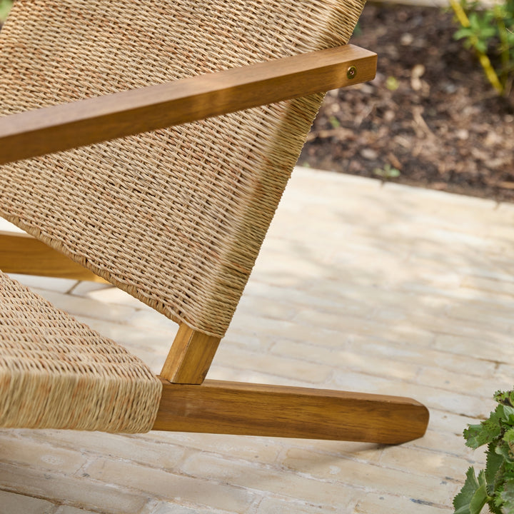 Close-up of a wooden outdoor lounge chair with woven all-weather wicker seat and back, shown on a light stone patio with garden mulch and plants in the background.