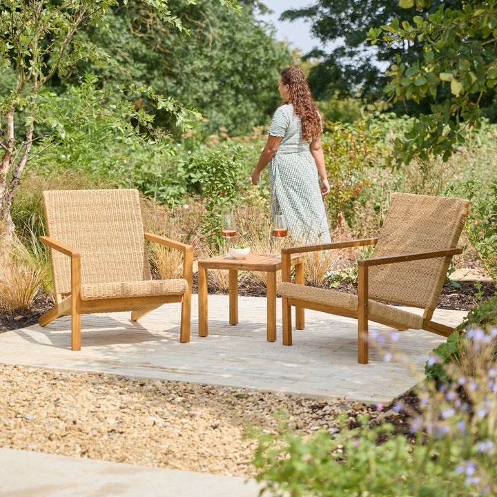 Two wooden patio armchairs casually arranged around a small matching wooden side table on a stone patio. A woman in a light dress walks in the background among green garden plants and trees, showing how large and generous the chairs are.