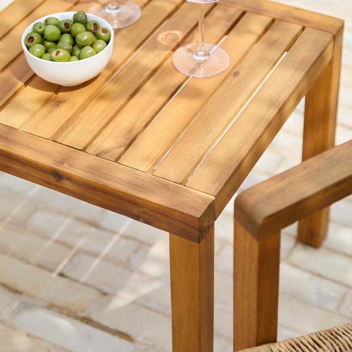 A close-up of a wooden outdoor table with a slatted top and matching wooden chair arm, styled with a bowl of green olives and wine glasses on a paved patio.