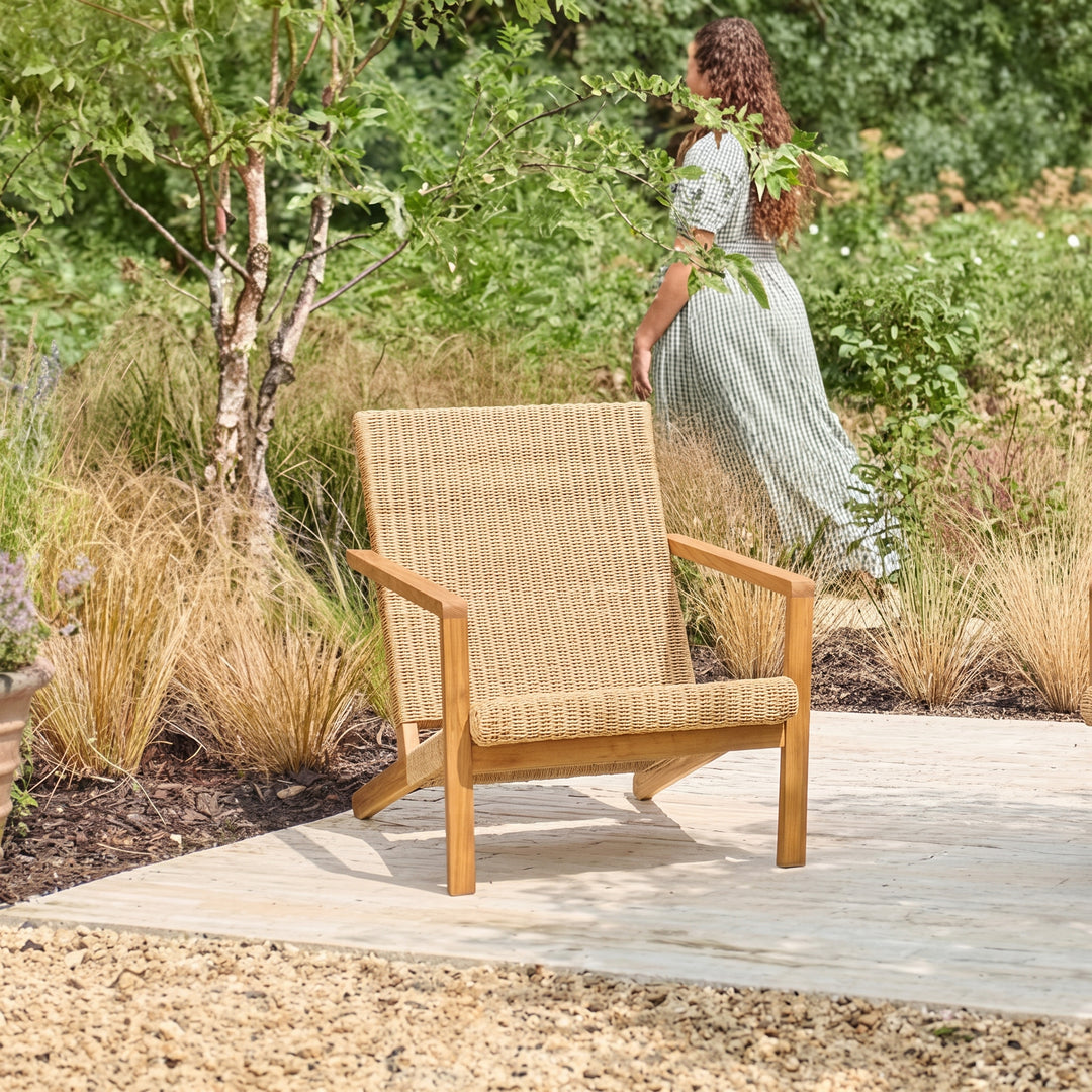 A modern outdoor lounge chair with a woven all-weather wicker seat and backrest and acacia wooden arms and legs, set on a stone patio in a garden with a woman walking in the background.