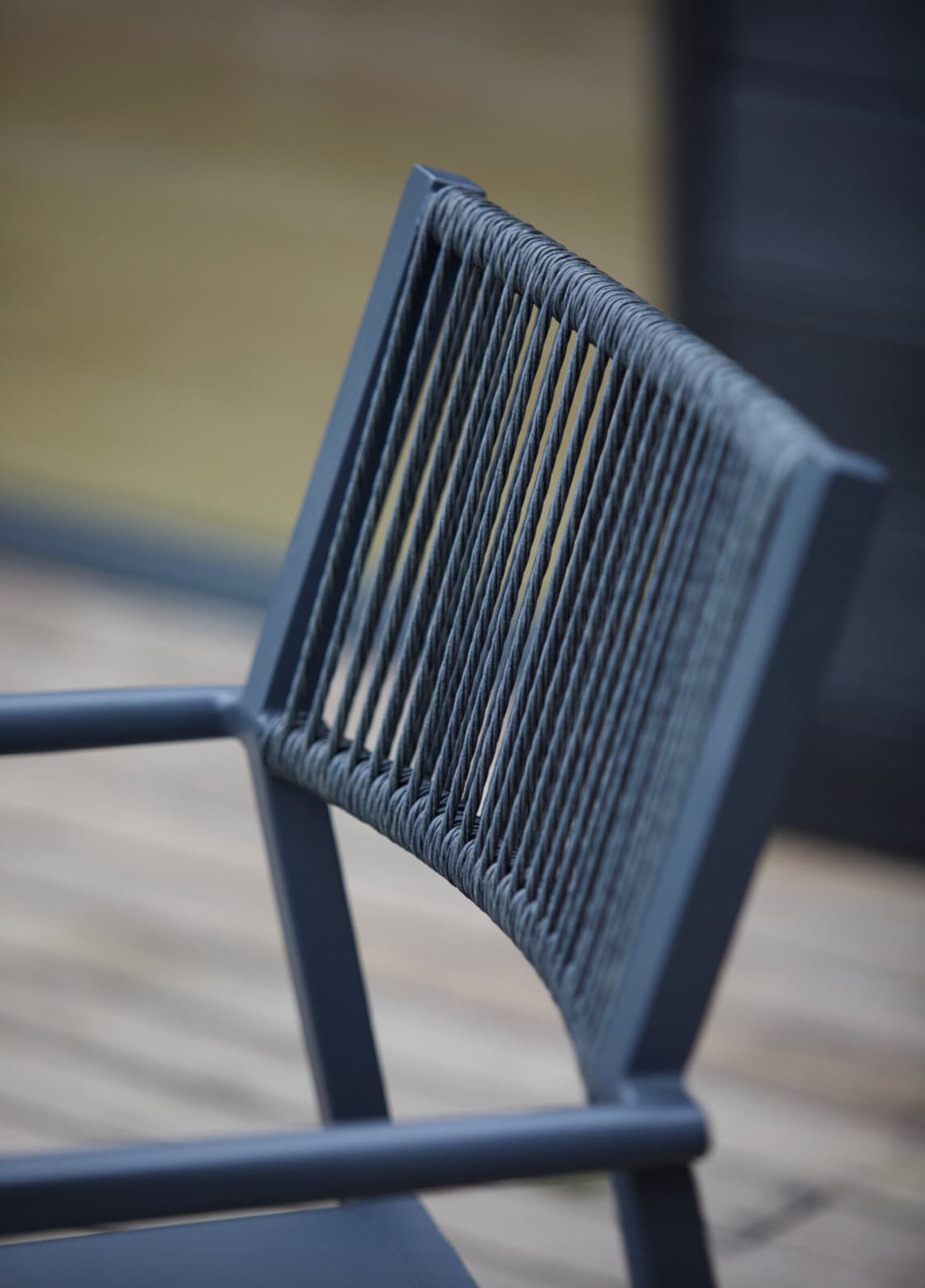 A close-up of a modern Richmond outdoor chair with a dark metal frame and a woven rope backrest. The tightly strung rope detailing creates a sleek, textured look. The chair is set on a wooden deck, with a blurred background suggesting an outdoor patio or garden setting.