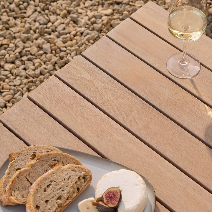 Repton Classic Wooden Garden Coffee Table with a slatted design, part of the Repton Corner Garden Furniture Set. Styled on a stone patio with two glasses of white wine and a bowl of snacks, this weather-resistant outdoor table complements modern patio and garden seating areas.