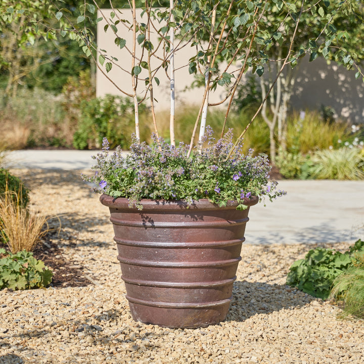 A large ribbed brown planter filled with green foliage and small purple flowers, placed outdoors on a gravel path in a landscaped garden.