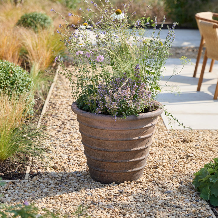A large ribbed terracotta planter filled with mixed wildflowers and green foliage, placed on a gravel path in a garden setting.