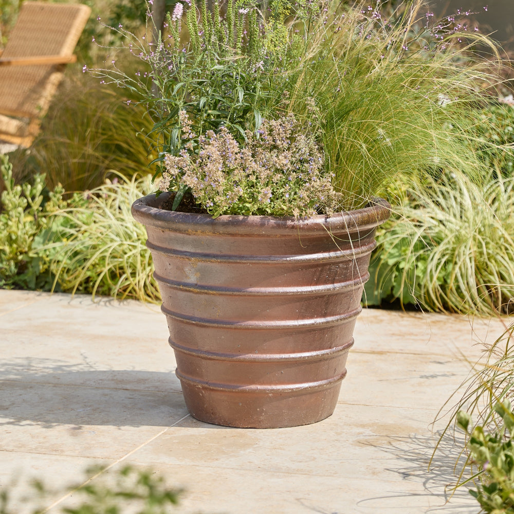 A large ribbed terracotta planter filled with ornamental grasses and small flowering plants, placed on a stone patio with garden foliage in the background.