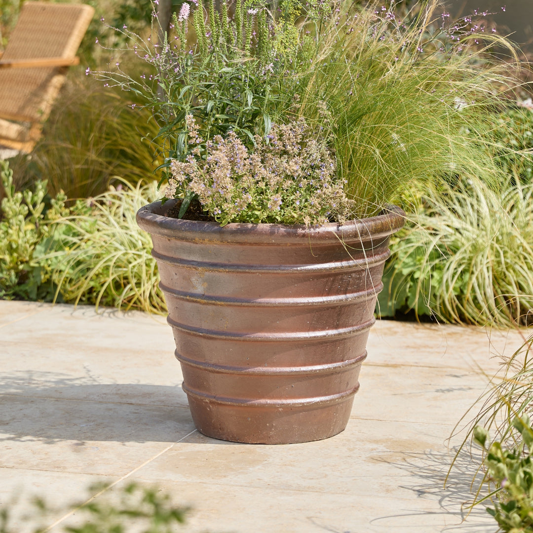 A large ribbed terracotta planter filled with ornamental grasses and small flowering plants, placed on a stone patio with garden foliage in the background.