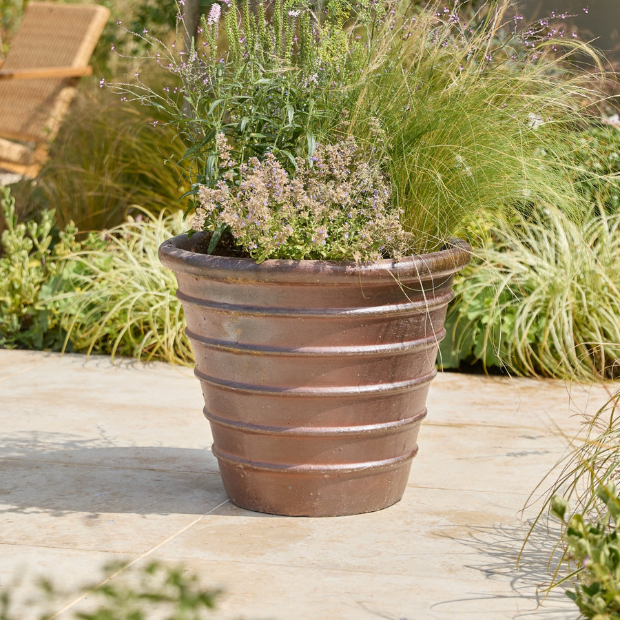 A large ribbed terracotta planter filled with ornamental grasses and small flowering plants, placed on a stone patio with garden foliage in the background.