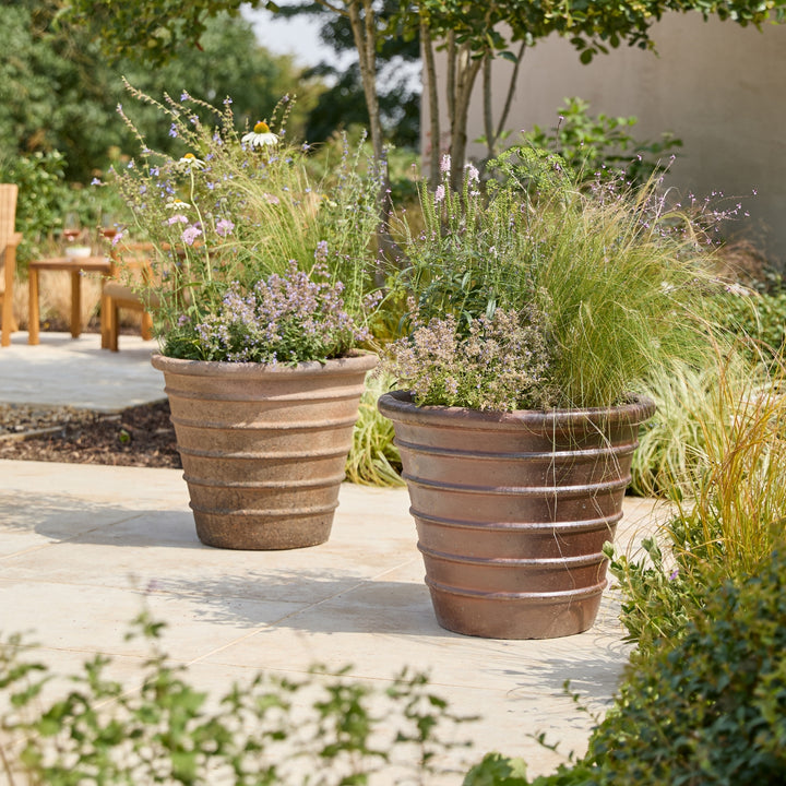Two large ribbed terracotta-colored planter pots filled with ornamental grass and small purple flowers, placed on a light stone patio in a landscaped garden setting.