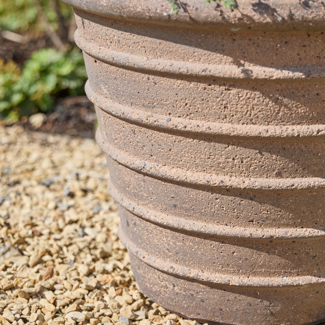 A close-up of a ribbed terracotta-colored plastic or concrete planter pot sitting on gravel outdoors.
